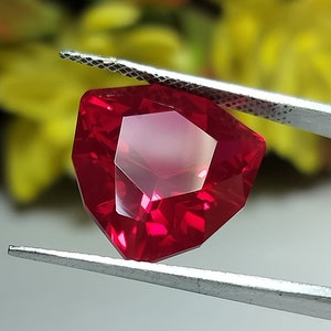 Puede incluir: Una piedra preciosa de color rojo intenso con forma octogonal facetada. La piedra preciosa está sujeta por unas pinzas.