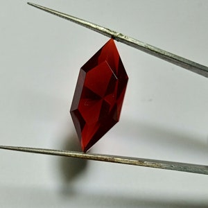 Op de afbeelding: Een dieprode edelsteen met een gefacetteerde, achthoekige vorm, vastgehouden door een pincet.