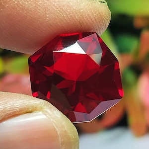 May include: A deep red octagonal gemstone with a faceted cut. The gemstone is held in a hand.