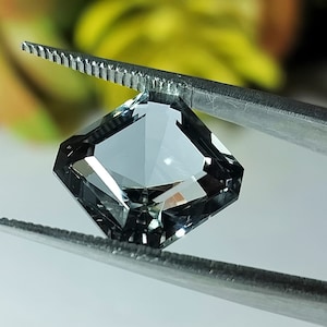 Könnte beinhalten: Ein klarer, facettierter Edelstein mit quadratischer Form, gehalten von einer Pinzette. Der Edelstein hat eine hellgraue Farbe und ist in eine rechteckige Form geschnitten.