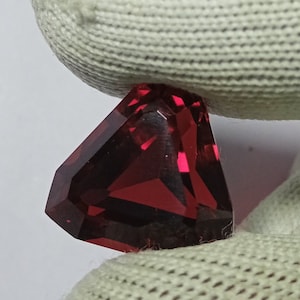 Pode incluir: Uma gema facetada, em forma triangular, com um tom vermelho profundo. O corte da gema revela múltiplas superfícies refletoras, criando um jogo de luzes. A gema é segurada contra um fundo branco.