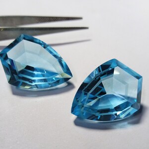 Könnte beinhalten: Zwei hellblaue Topas-Edelsteine, in Tropfenform geschliffen, mit facettiertem Design. Die Edelsteine liegen auf einer weißen Oberfläche.