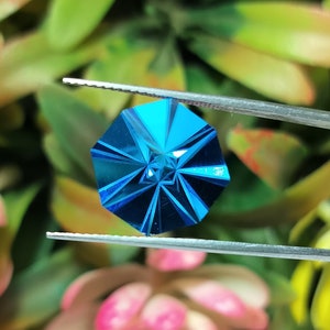 May include: A faceted blue gemstone with a star-shaped design in the centre. The gemstone is held by tweezers against a green and pink background.