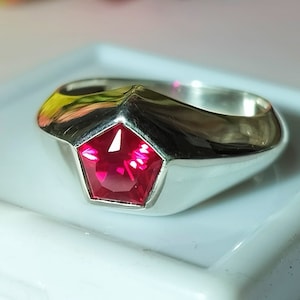Könnte beinhalten: Ein silberner Ring mit einem fünfeckigen, leuchtend roten Edelstein. Der Ring hat eine polierte, reflektierende Oberfläche und ein breites Band. Der Edelstein ist facettiert und fängt das Licht ein, wodurch ein funkelnder Effekt entsteht.