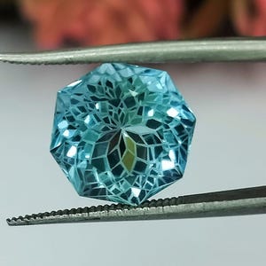 May include: A light blue gemstone with a faceted, starburst design. The gemstone is held by tweezers.