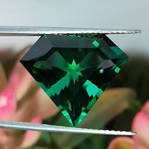 May include: A deep green gemstone with a star-shaped pattern in the centre, held by tweezers against a blurred floral background.