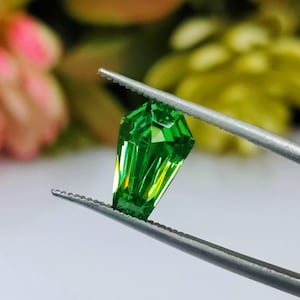 May include: A vibrant green gemstone with a pointed, faceted shape, held by tweezers against a blurred background.
