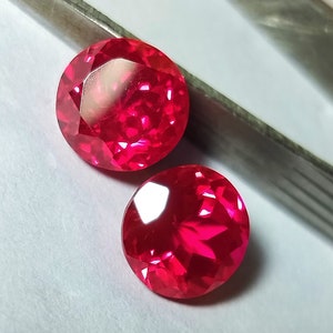 May include: Two round, red gemstones with a faceted cut. The gemstones are on a white surface.