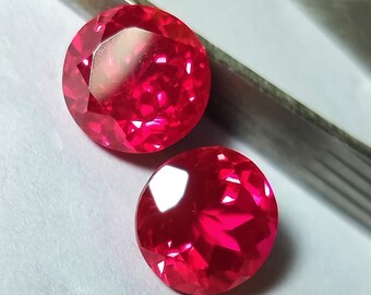 May include: Two round, red gemstones with a faceted cut. The gemstones are on a white surface.