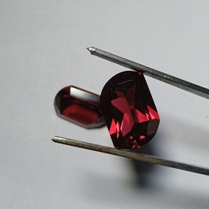 May include: Two red gemstones, one held by tweezers, with a faceted oval shape.