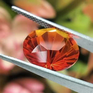 May include: A bright orange gemstone with a faceted cut, held in tweezers against a blurred background.