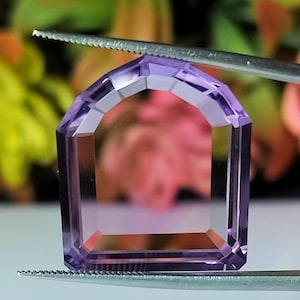 Puede incluir: Una gema de amatista facetada en forma de arco, sujeta con pinzas. La gema es de color púrpura claro, con una base rectangular y una parte superior arqueada. Las facetas crean un juego de luces y sombras.