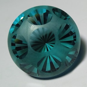 Può includere: Un fermacarte in vetro blu turchese con un design geometrico. Il fermacarte ha una superficie liscia e arrotondata ed è decorato con un motivo di linee radiali.