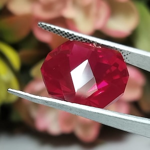 May include: A faceted, octagonal ruby gemstone held by silver tweezers. The ruby is a deep, vibrant red with multiple reflective surfaces. The background is a soft, blurred mix of pink and green.