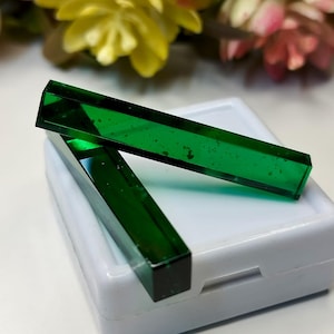 Puede incluir: Dos piedras preciosas rectangulares de color verde esmeralda en una caja blanca. Las piedras preciosas son translúcidas y tienen un ligero brillo.