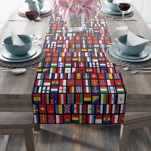 May include: A rectangular table runner featuring a pattern of various national flags on a black background. The runner is placed on a wooden table set for a meal, with plates, bowls, glasses, and silverware.