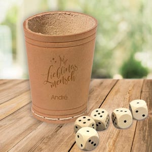 Könnte beinhalten: Ein brauner Leder-Würfelbecher mit dem eingravierten Text "My Lieblingsmensch Andre". Fünf Holzwürfel mit schwarzen Punkten liegen auf einer Holzoberfläche verstreut.