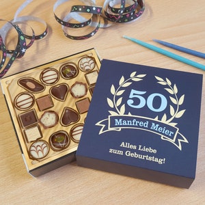 Könnte beinhalten: Eine Schachtel Pralinen mit goldfarbenem Innenraum neben einer marineblauen Geschenkbox. Die Geschenkbox trägt die Zahl 50, den Namen "Manfred Meier" und den Text "Alles Liebe zum Geburtstag!" in Gold.