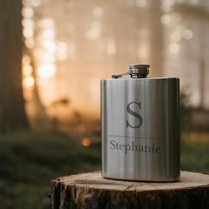 Könnte beinhalten: Eine silberfarbene Hüftflasche mit Klappverschluss, graviert mit dem Buchstaben "S" und dem Namen "Stephanie". Die Flasche hat eine abgerundete rechteckige Form und steht auf einem Baumstumpf, mit verschwommenem Waldbild im Hintergrund.
