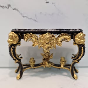 Puede incluir: Una consola de estilo barroco negra y dorada con una tapa de mármol. La mesa tiene tallas y detalles intrincados, incluyendo querubines y motivos florales.