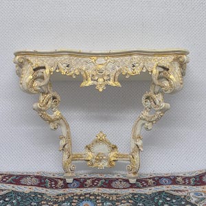 Puede incluir: Una consola de pared con acabado en blanco roto y tallas ornamentadas pintadas en oro. La pieza muestra intrincados motivos de volutas, elementos florales y una pieza decorativa central que conecta las dos patas curvas bajo el estante.