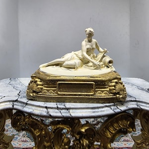 Puede incluir: Una figura de color crema de una silueta reclinada envuelta en tela, descansando junto a un pequeño recipiente. La escultura está montada sobre una base rectangular dorada y ornamentada con motivos de conchas en las esquinas y una placa.