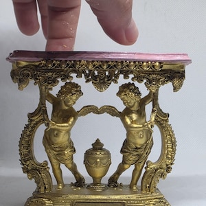 以下が含まれることがあります： ピンクの天板を持つ金色のミニチュアテーブル。フレームを支える2体の天使のフィギュアと中央の壺が特徴です。台座は長方形で装飾が施されており、下部にはREINAMINIATURESという文字が印刷されています。