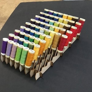 Op de afbeelding: Een houten garenrek met een trapsgewijs ontwerp bevat een verscheidenheid aan kleurrijke garenklossen. De klossen zijn in rijen gerangschikt, waarbij de kleuren overgaan van donkerblauw naar rood.