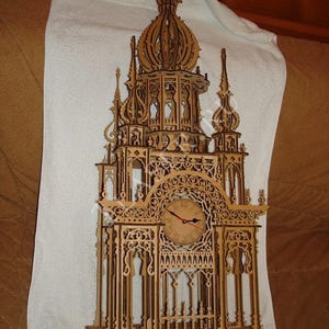 Può includere: Una torre dell'orologio in legno con intagli intricati e un quadrante dell'orologio con numeri romani. La torre dell'orologio è in legno marrone chiaro e ha un'alta guglia appuntita.