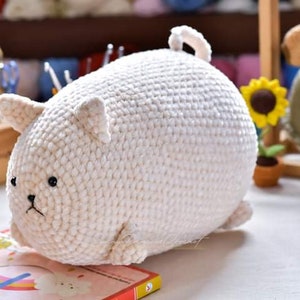 May include: A white crocheted stuffed animal shaped like a cat. The cat has black eyes and a small black nose. The cat is lying on a book with a rainbow design.