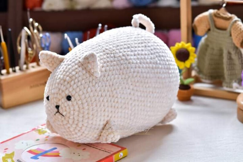 May include: A white crocheted stuffed animal shaped like a cat. The cat has black eyes and a small black nose. The cat is lying on a book with a rainbow design.