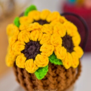 Puede incluir: Una maceta de girasol de ganchillo hecha con hilo marr&oacute;n, amarillo y verde. La maceta est&aacute; sentada sobre una superficie de madera. Sunny Princess Crochet est&aacute; escrito en la parte inferior de la maceta.