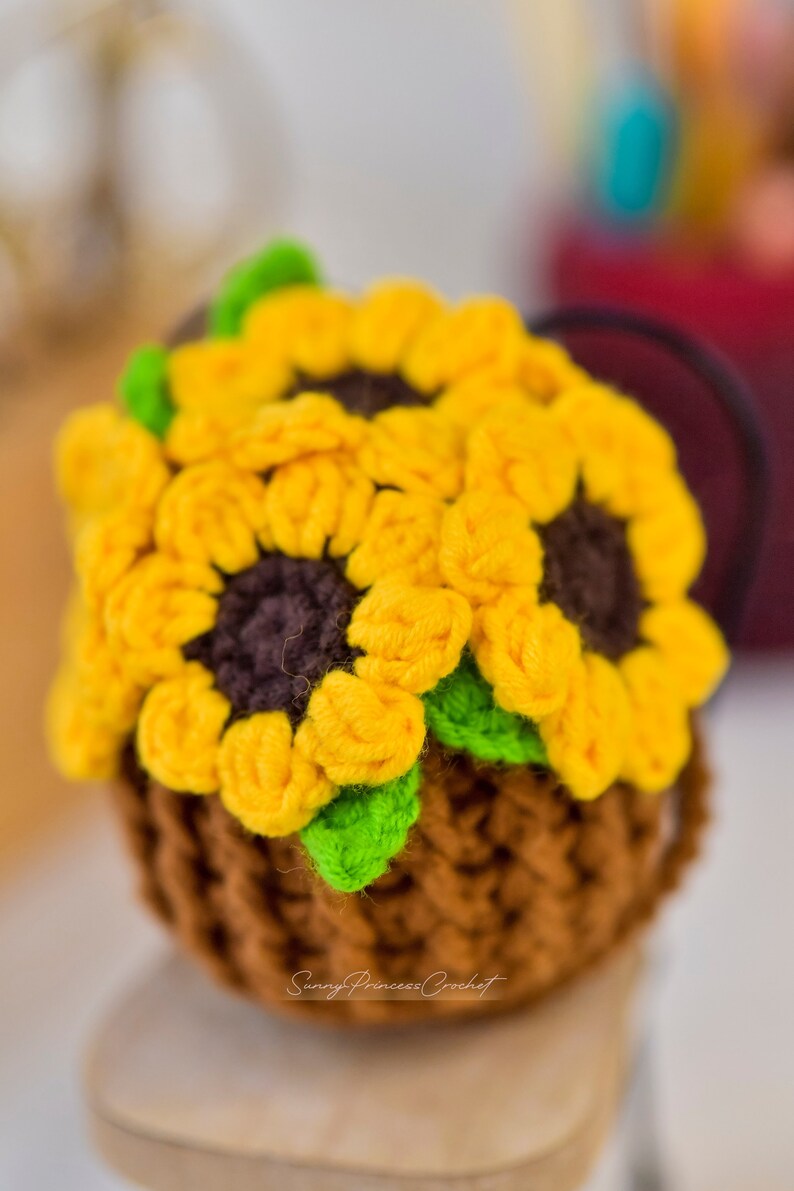 Puede incluir: Una maceta de girasol de ganchillo hecha con hilo marr&oacute;n, amarillo y verde. La maceta est&aacute; sentada sobre una superficie de madera. Sunny Princess Crochet est&aacute; escrito en la parte inferior de la maceta.