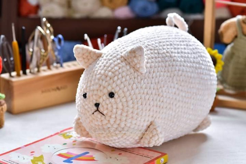 May include: A white crocheted stuffed animal shaped like a cat with black eyes. The cat is sitting on a book with a rainbow design.