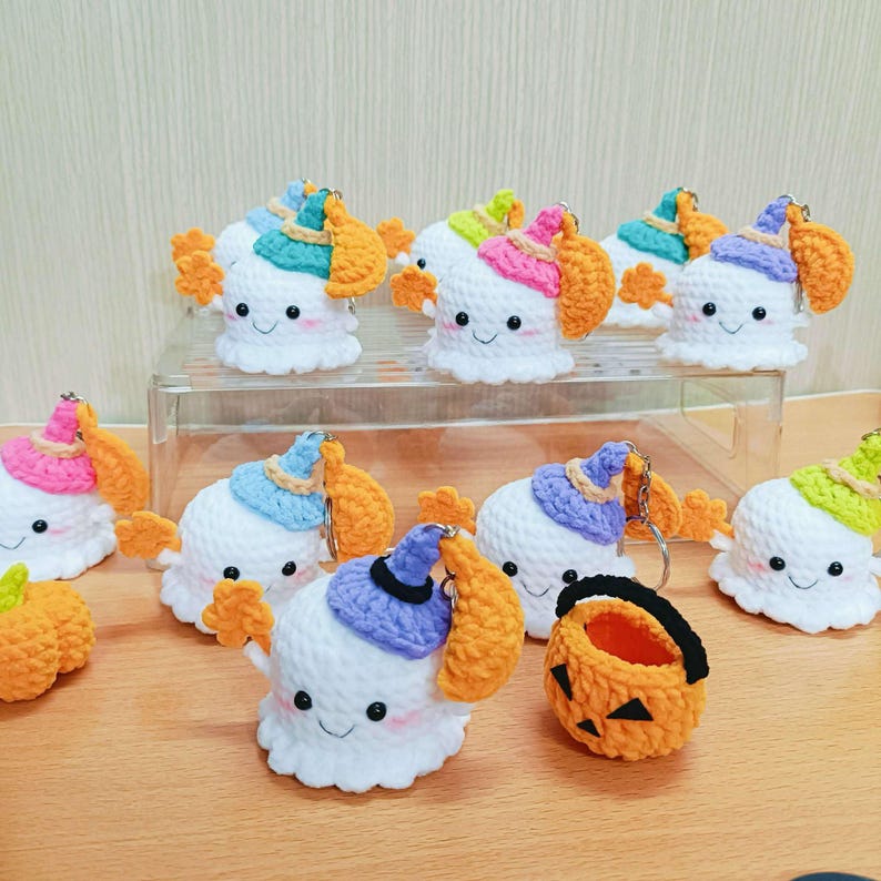 Puede incluir: Una colecci&oacute;n de llaveros de fantasmas de Halloween hechos a crochet con sombreros de diferentes colores y un cubo de calabaza hecho a crochet. Los fantasmas tienen cuerpos blancos, ojos negros y una boca sonriente. Los sombreros son morados, azules, verdes y rosas. El cubo de calabaza es naranja con una cara de calabaza negra.