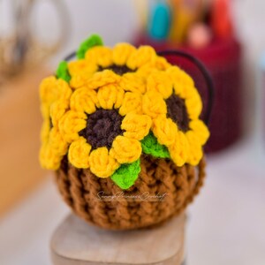Puede incluir: Una maceta de girasol de ganchillo hecha con hilo marr&oacute;n, amarillo y verde. La maceta est&aacute; sobre una superficie de madera.