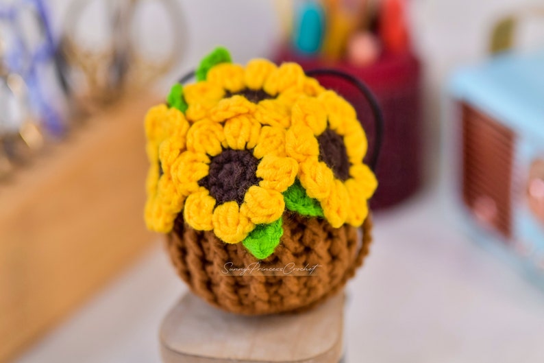 Puede incluir: Una maceta de girasol de ganchillo hecha con hilo marr&oacute;n, amarillo y verde. La maceta est&aacute; sobre una superficie de madera.