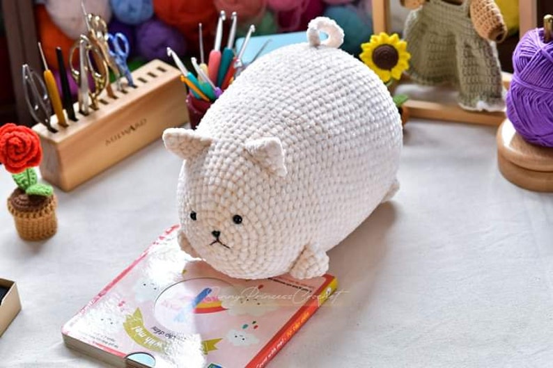 May include: A white crocheted plush toy shaped like a cat. The toy has a round body and small ears. It is sitting on a book with a rainbow design.