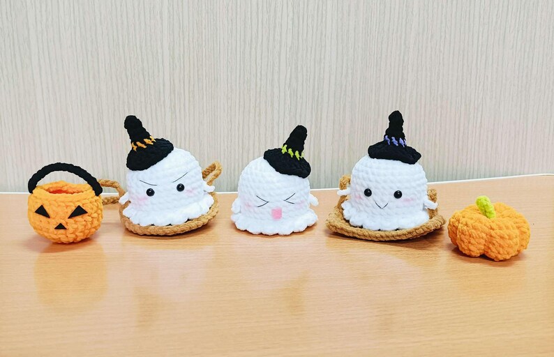 Puede incluir: Cuatro decoraciones de Halloween de ganchillo: tres fantasmas con sombreros de bruja y una peque&ntilde;a calabaza naranja. Los fantasmas tienen ojos negros y bocas sonrientes. La calabaza tiene un tallo verde.