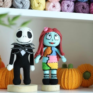 May include: Crocheted amigurumi dolls of Jack Skellington and Sally from the Nightmare Before Christmas film. The dolls are standing on wooden bases and are surrounded by orange pumpkins.