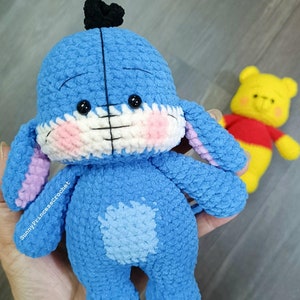 Puede incluir: Un juguete de peluche azul tejido a crochet que se asemeja a un personaje de dibujos animados con orejas grandes, una nariz negra y un lazo negro en la cabeza. El juguete tiene una boca cosida blanca y mejillas rosadas. El juguete está de pie sobre una mano.