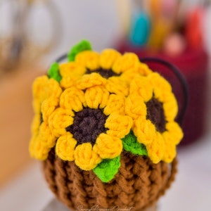 Puede incluir: Una maceta de girasol de ganchillo con tres girasoles amarillos y hojas verdes. La maceta es marr&oacute;n y tiene una superficie texturizada. La maceta est&aacute; sentada sobre una superficie de madera.