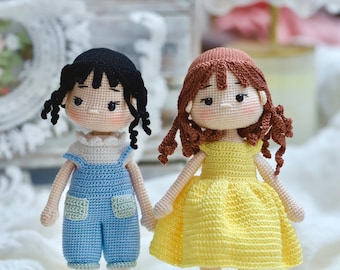 May include: Two handmade crochet dolls. One doll wears blue overalls and has black hair. The other doll wears a yellow dress and has brown hair. Both dolls are holding hands and have rosy cheeks. The dolls are standing on a white lace surface.