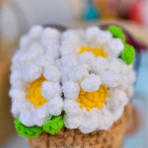 May include: A crocheted flower bouquet in a brown pot with green leaves. The flowers are white with yellow centers. The pot has the text "Sunny Princess Crochet" on the side.