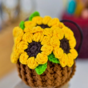 Puede incluir: Una maceta de girasol de ganchillo con tres girasoles amarillos y hojas verdes. La maceta es marr&oacute;n y tiene una superficie texturizada. La maceta est&aacute; sentada sobre una superficie de madera.