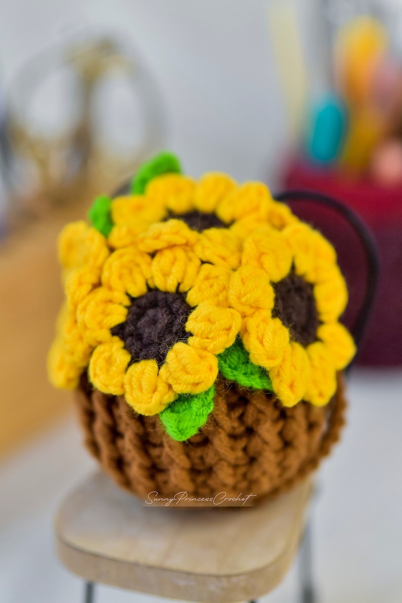 Puede incluir: Una maceta de girasol de ganchillo con tres girasoles amarillos y hojas verdes. La maceta es marr&oacute;n y tiene una superficie texturizada. La maceta est&aacute; sentada sobre una superficie de madera.