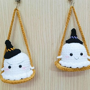 Puede incluir: Dos adornos de fantasmas de crochet con sombreros de bruja negros colgando de una cuerda marr&oacute;n. Los fantasmas tienen cuerpos de lana blanca y ojos negros. Un fantasma tiene una cara fruncida y el otro tiene una cara sonriente.