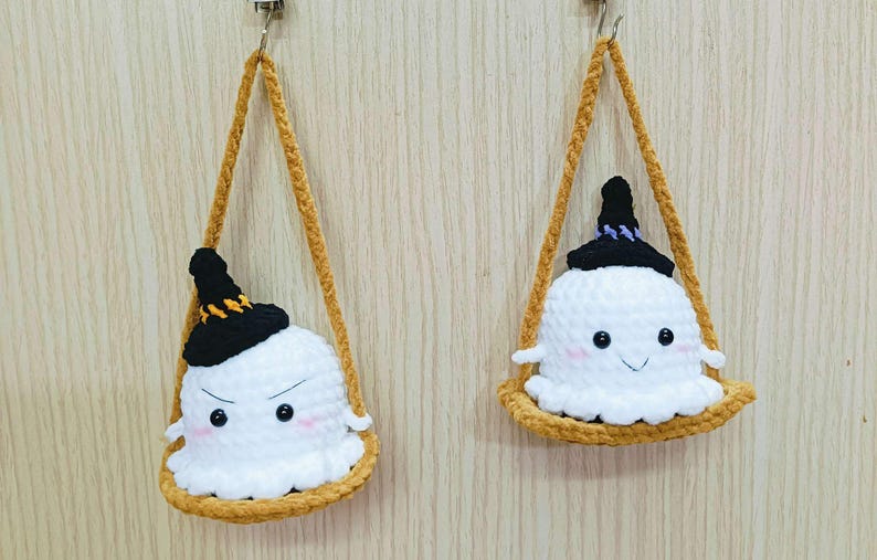 Puede incluir: Dos adornos de fantasmas de crochet con sombreros de bruja negros colgando de una cuerda marr&oacute;n. Los fantasmas tienen cuerpos de lana blanca y ojos negros. Un fantasma tiene una cara fruncida y el otro tiene una cara sonriente.
