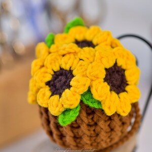 Puede incluir: Una maceta de girasol de ganchillo con hilo marr&oacute;n para la maceta e hilo amarillo y verde para los girasoles. La maceta est&aacute; sentada en un peque&ntilde;o taburete de madera.