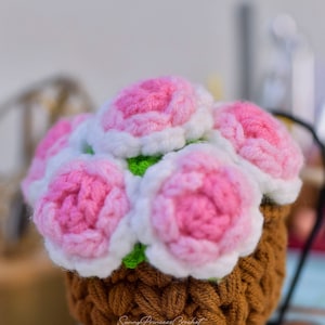 Könnte beinhalten: Eine gehäkelte Blumenarrangements in einem braunen Topf. Die Blumen sind rosa und weiß mit grünen Blättern. Der Topf hat eine gewebte Textur. Der Text "Sunny Princess Crochet" ist auf dem Topf sichtbar.